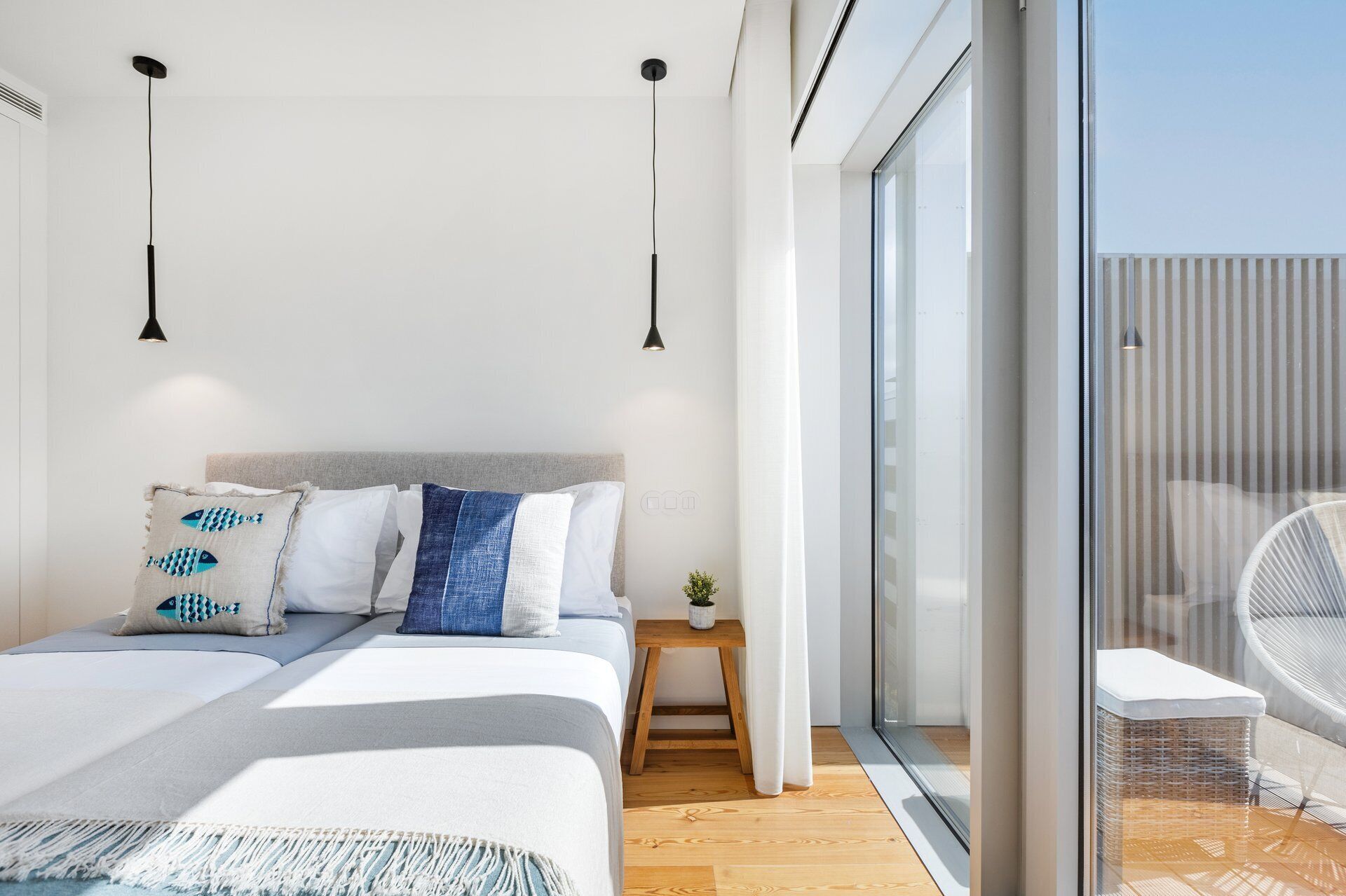 A modern bedroom with a large bed, two pendant lights, a small wooden nightstand, and a view of the outside through a sliding glass door.