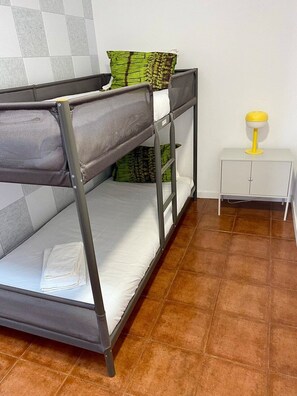 A bunk bed with a white mattress and a grey metal frame. There is a small white nightstand with a yellow lamp beside it. - Live Well, Beach Guest Home with Pool (with A/C) (Vila Nova de Gaia)