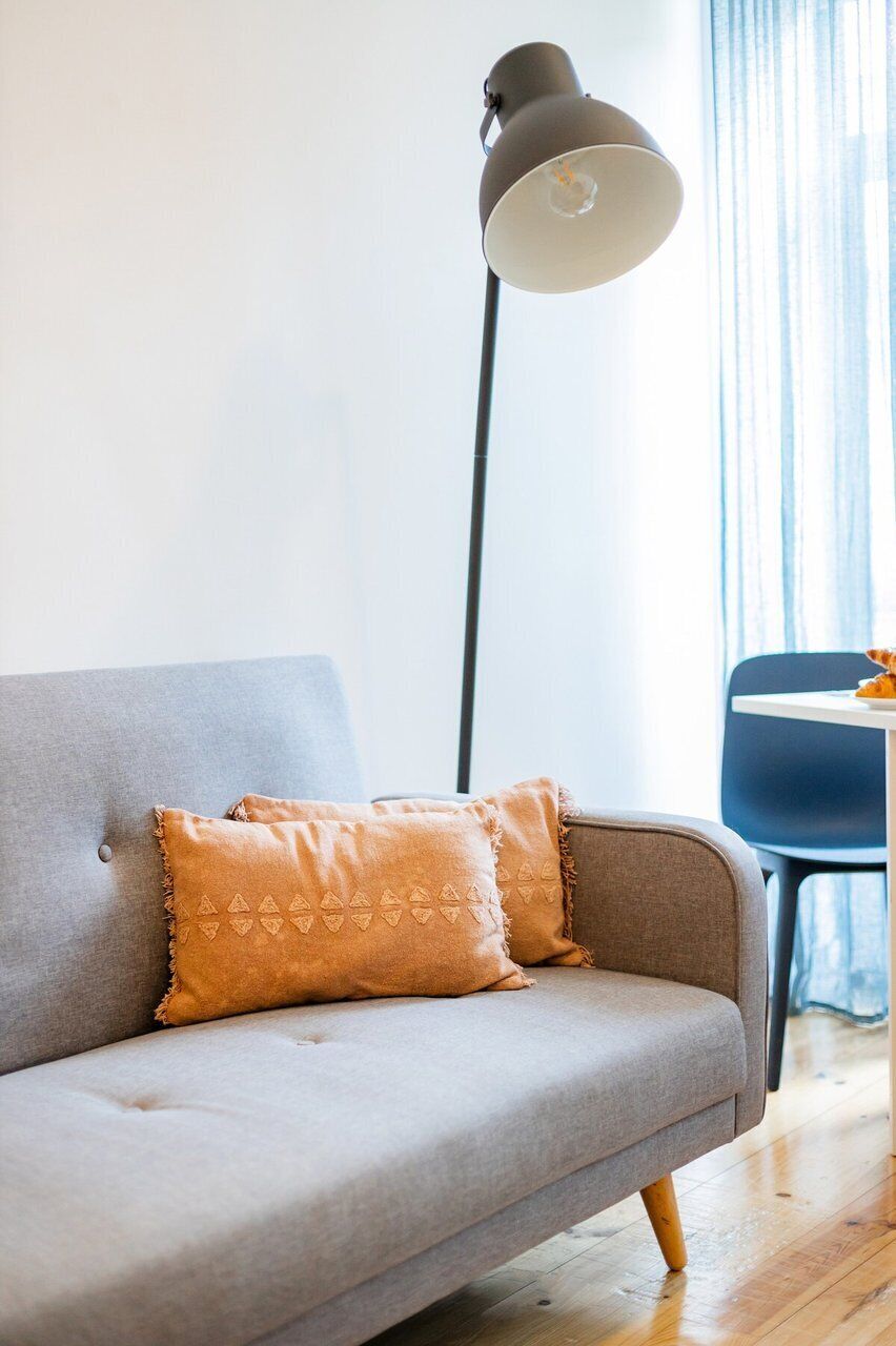 Un sofá gris con dos cojines estampados, una lámpara de pie alta y una silla azul en una habitación con suelo de madera.