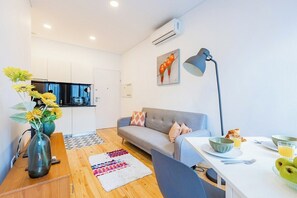 Uma sala de estar moderna com um sofá, uma mesa de jantar com itens de café da manhã, um vaso com flores e uma cozinha ao fundo.