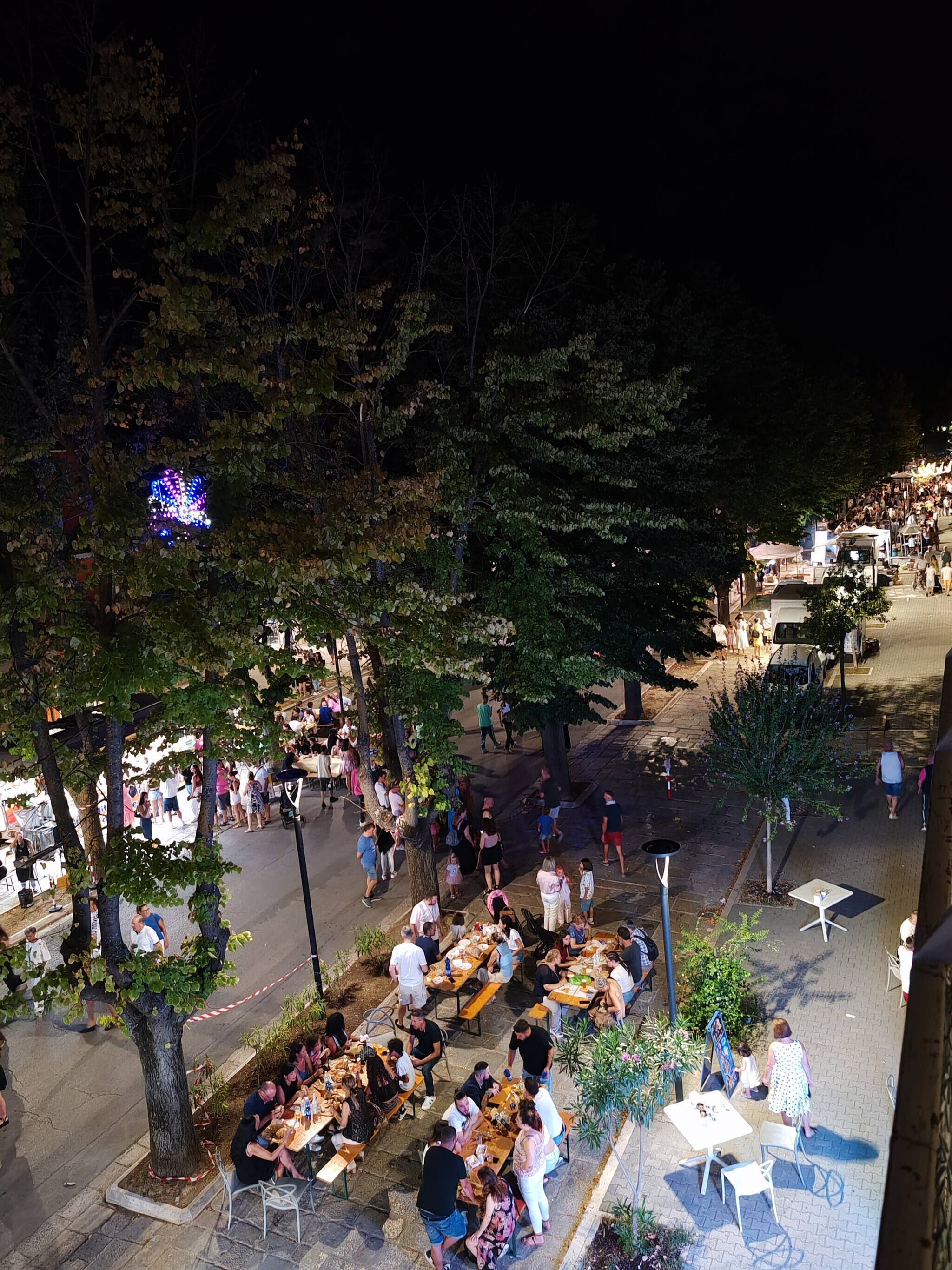 An outdoor dining area with tables and chairs, surrounded by trees and a street.