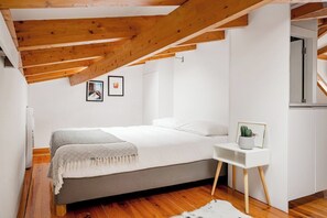 A bedroom with a bed, wooden beams, white walls, a small bedside table with a plant, and framed pictures on the wall. - Arriaga River Views II (Lisbon)