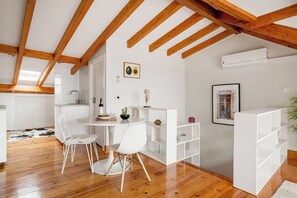A modern kitchen with white cabinetry, a dining area with a round table and chairs, wooden beams on the ceiling, and a white shelving unit. - Arriaga River Views II (Lisbon)