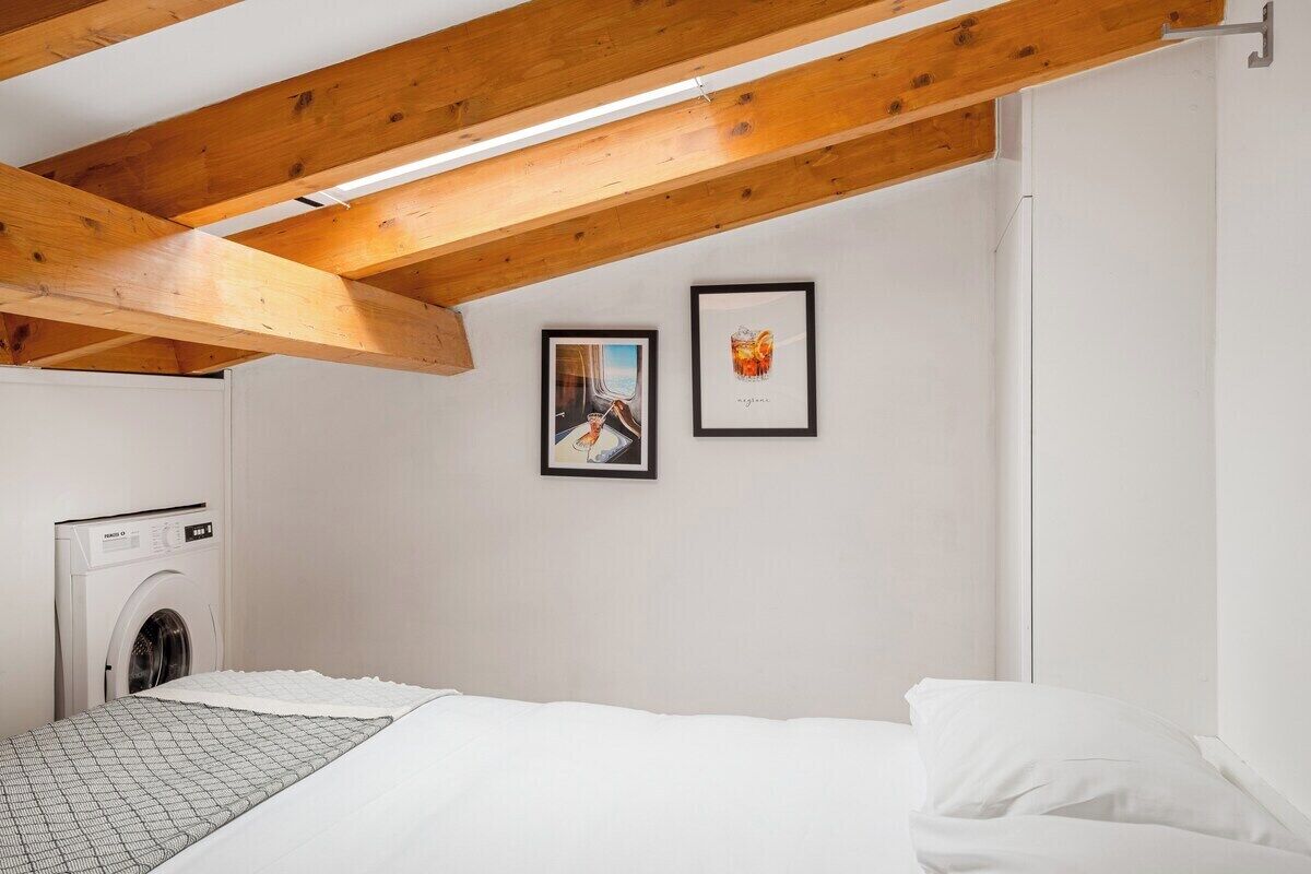 A bedroom with a bed, a washing machine, wooden beams, and two framed pictures on the wall.
