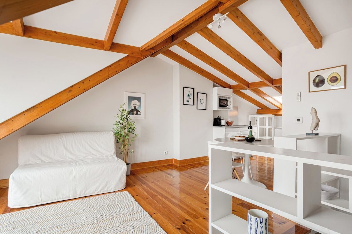 Un dormitorio tipo loft con techo de madera, muebles blancos, un sofá blanco, una planta y una zona de cocina con armarios blancos.