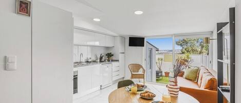 A modern kitchen with white cabinets, a dining table with breakfast items, and a sofa with a view to the outdoors.