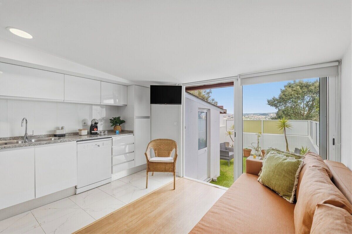 A modern kitchen with white cabinets, a dining area with a wicker chair, and a balcony with a view.