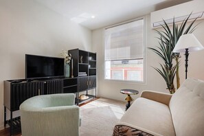 Ein Wohnzimmer mit Fernseher, schwarzem Regalelement, einem hellgrünen Sessel, einem beigefarbenen Sofa, einem kleinen runden Tisch und einem großen Fenster mit Jalousien.