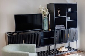 Ein modernes Wohnzimmer mit einem Flachbildfernseher, einem hellen Sessel, einem dunklen Holzkonsole und einer schwarzen Regaleinheit mit Dekorationsobjekten.