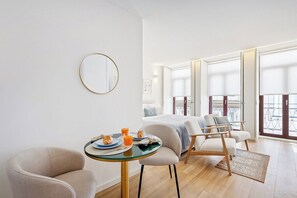 Una habitación de hotel moderna con una cama, dos sillas, una mesa redonda con desayuno y ventanas amplias.