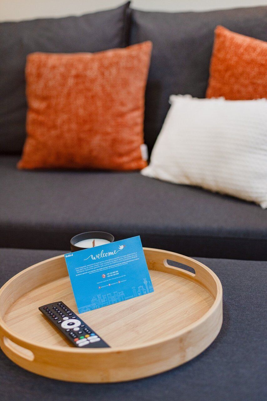 A wooden tray with a remote control and a blue card on a dark sofa with orange and white pillows.
