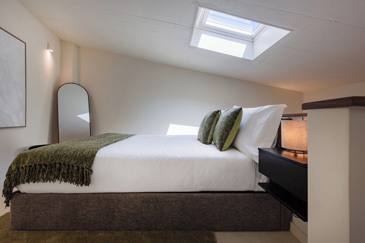 A bedroom with a bed, a bedside table, a lamp, a mirror, and a skylight.