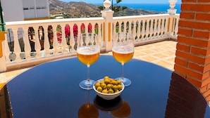 Een tafel met twee bierglazen en een schaal olijven, op een balkon met uitzicht op zee en heuvels.