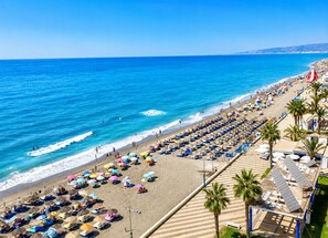 Une plage bordée de rangées de transats et de parasols, une mer d’un bleu limpide et une côte lointaine.