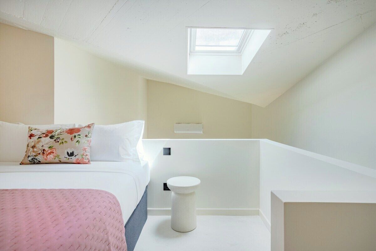 Un dormitorio con tragaluces, una cama con almohadones florales, una mesita de noche y un taburete blanco pequeño.