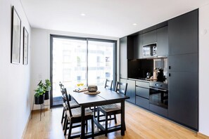 A modern kitchen with dark cabinetry, a dining table with chairs, and a large window providing a view of the city. - Camões Downtown with Parking (Porto)