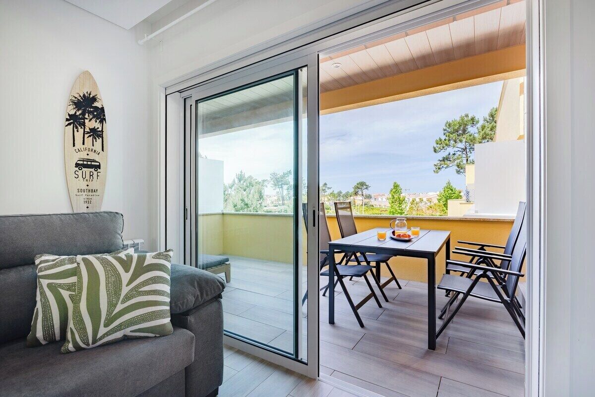 A modern living room with a gray sofa, a surfboard decoration, and a sliding glass door leading to an outdoor patio with a table and chairs.