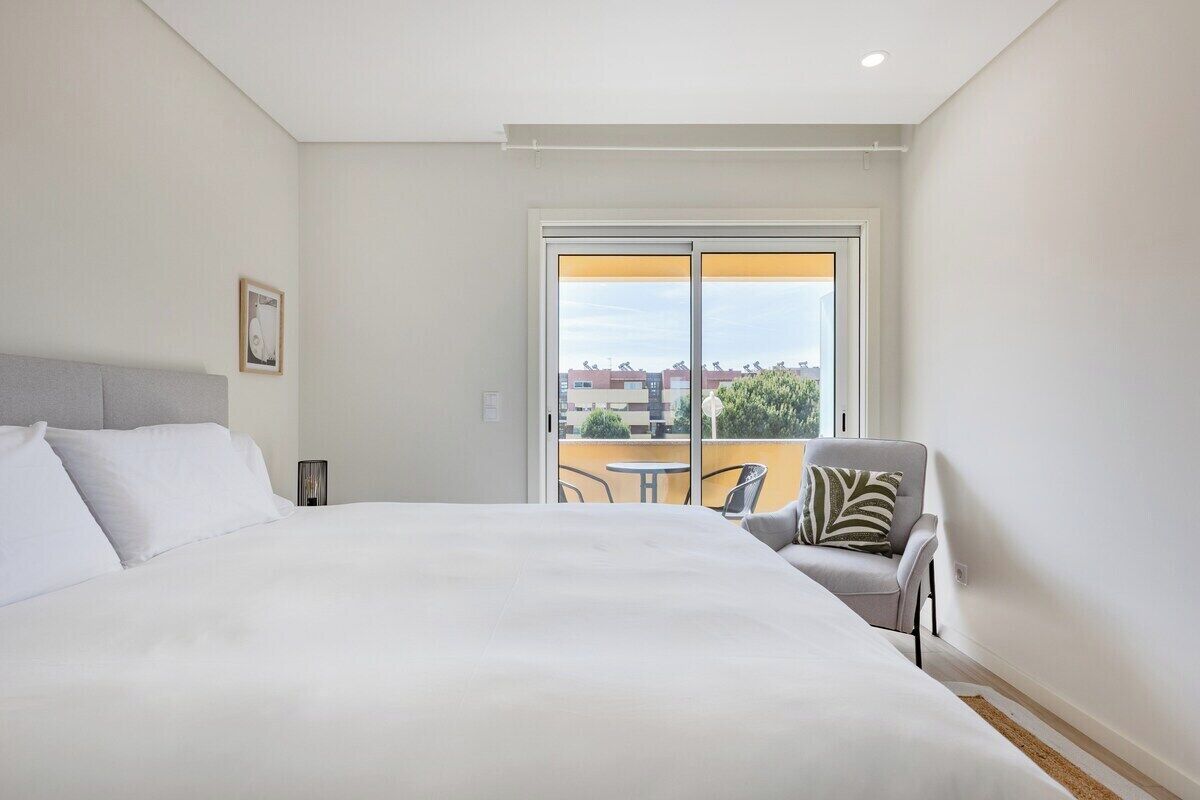 A hotel room with a large bed, a chair, a small table, and a balcony with a view of buildings.