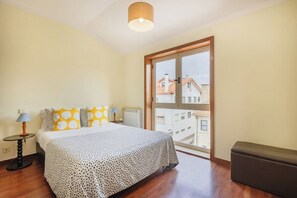 Un dormitorio con una cama, una mesita de noche, una lámpara, un banco y un amplio ventanal con vistas a edificios.