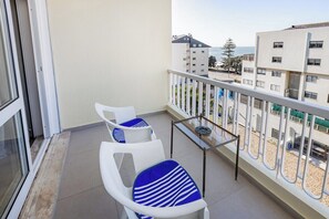 Ein Balkon mit weißen Stühlen und einem kleinen Tisch, mit Blick auf ein Gebäude und einen Strand.