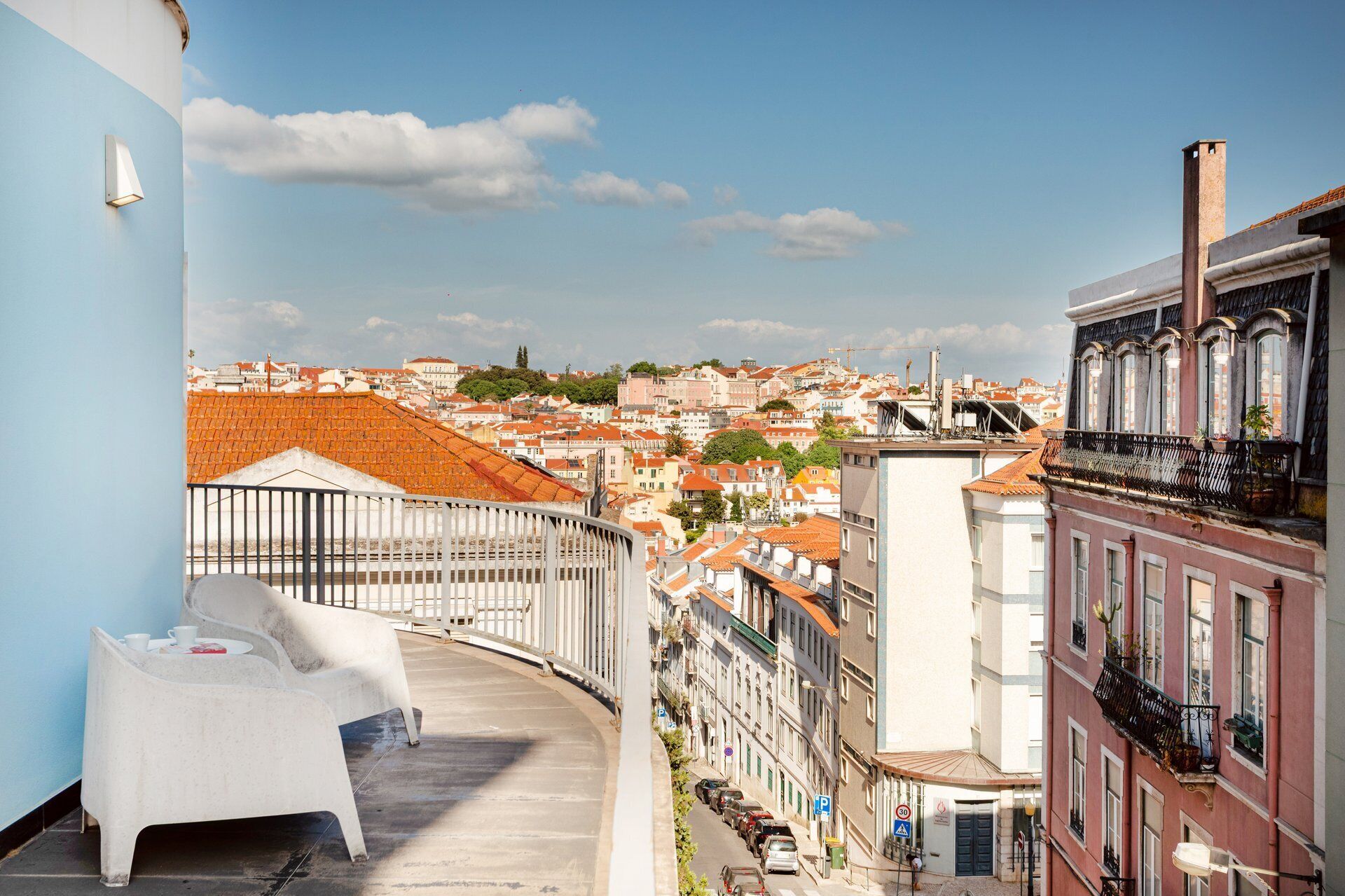 A balcony with a white chair, a railing, and a view of a cityscape with red-roofed buildings.