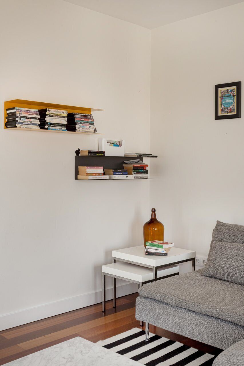 A living room with a grey sofa, a white coffee table with books, a small white side table, and two shelves with books and decorative items.