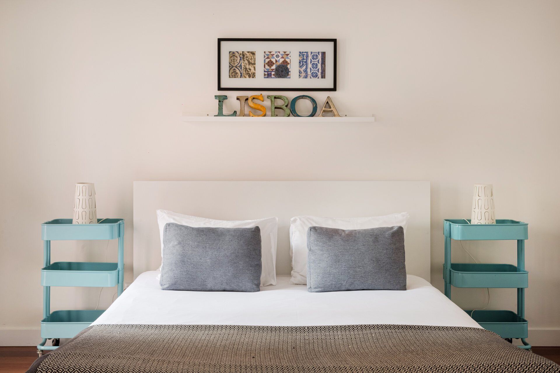 Een bed met twee kussens, een lijst met een afbeelding erboven en een plank waarop de letters 'LISBOA' staan.
