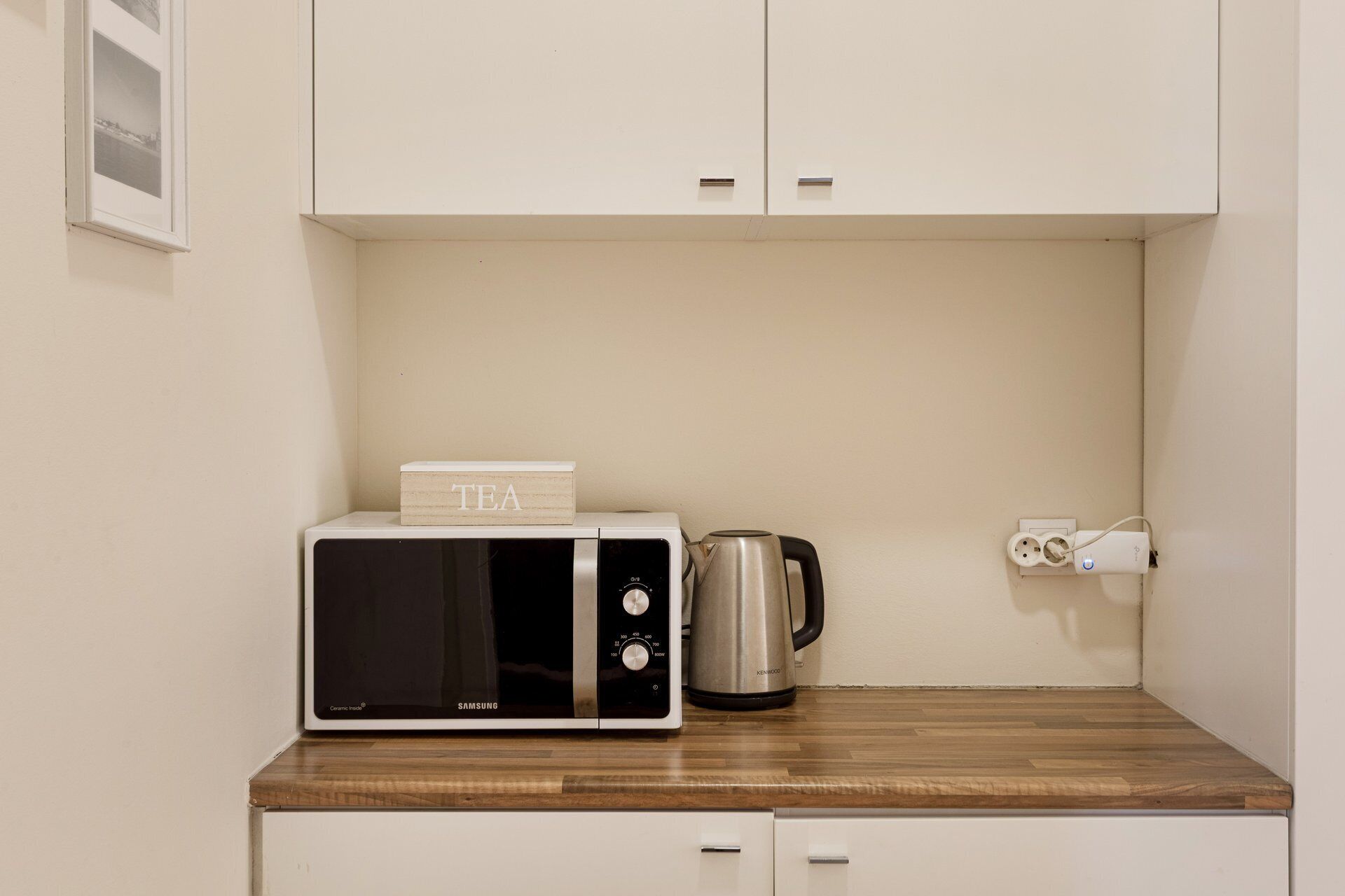 A kitchen with a microwave, a kettle, and a power outlet on a wooden countertop.