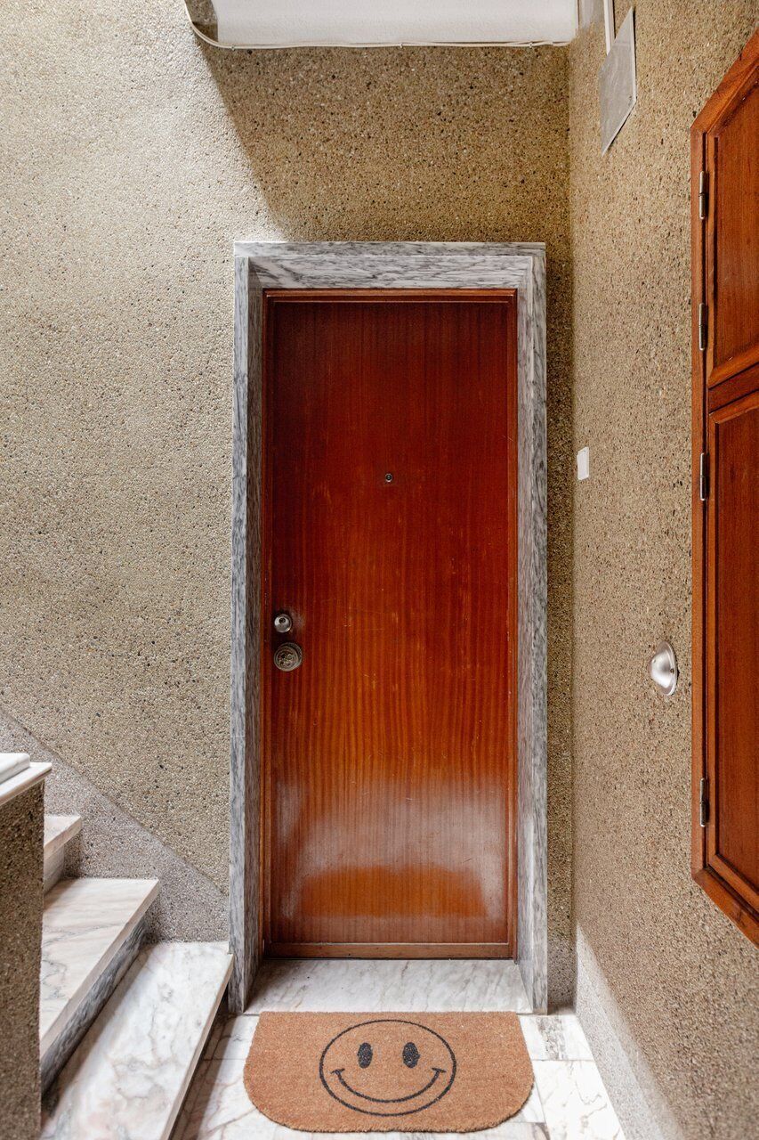 A wooden door with a metal handle, a marble threshold, and a smiley face mat on the floor.