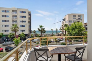 A balcony with a table and chairs, overlooking a street with parked cars and buildings, and a view of the beach and ocean. - Casa Del Sol SeaSide (Durrës)