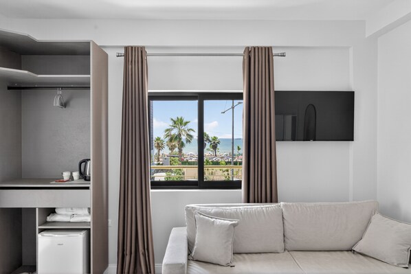 A modern hotel room with a sofa, a small kitchenette, a window with curtains, and a view of the beach.
