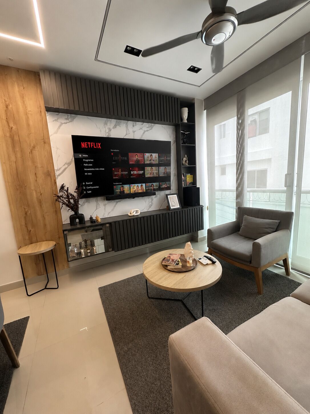 A modern living room with a flat-screen TV displaying a Netflix interface, a ceiling fan, and a large window offering a view of buildings.