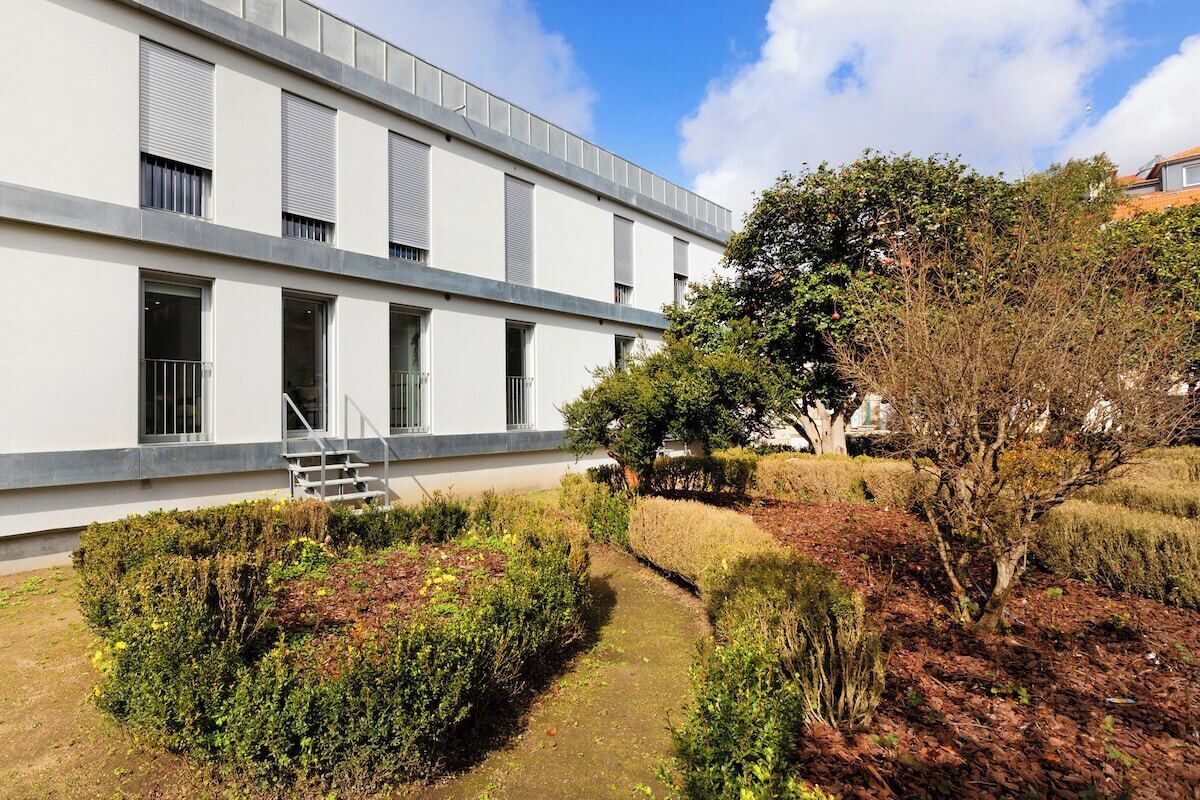 Um edifício moderno com telhado plano, grandes janelas e um jardim bem cuidado com sebes e árvores.