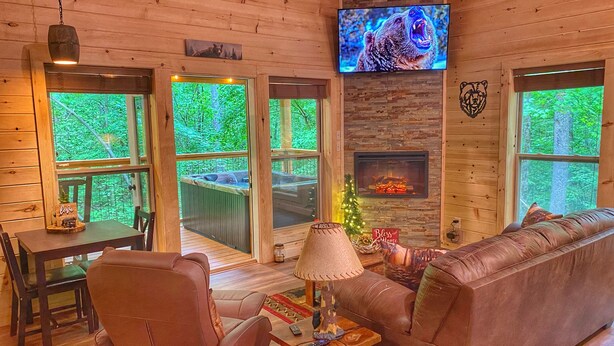 A cozy cabin interior with a fireplace, a television mounted on the wall, a leather sofa, and a dining area with a table and chairs. - NEW LISTING~Quiet~cozy~clean~fast WIFI, Dollywood, hotub/FP #9 (Sevierville)