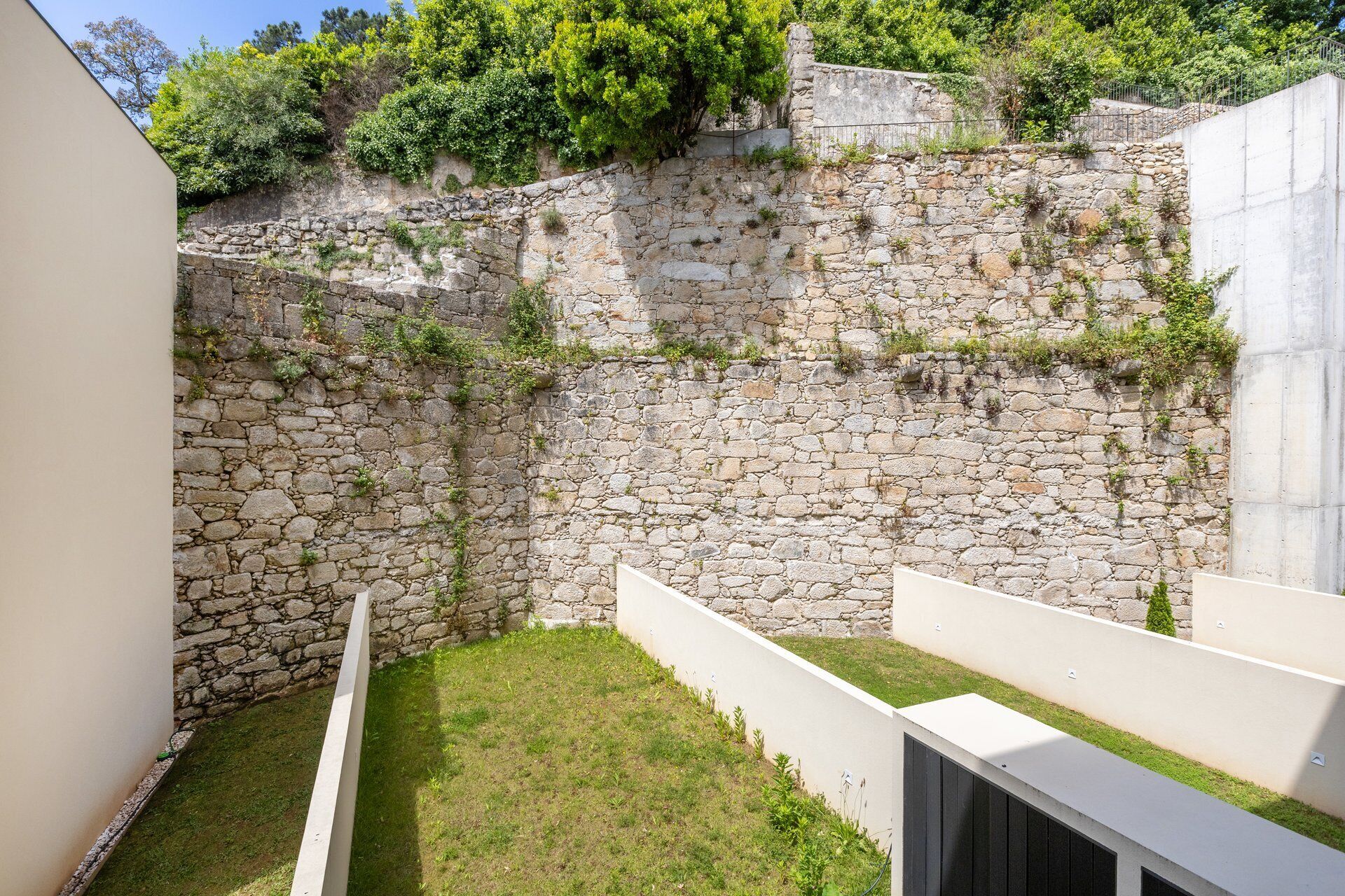 A stone wall with a grassy area in front, a white structure to the left, and a metal fence on top.