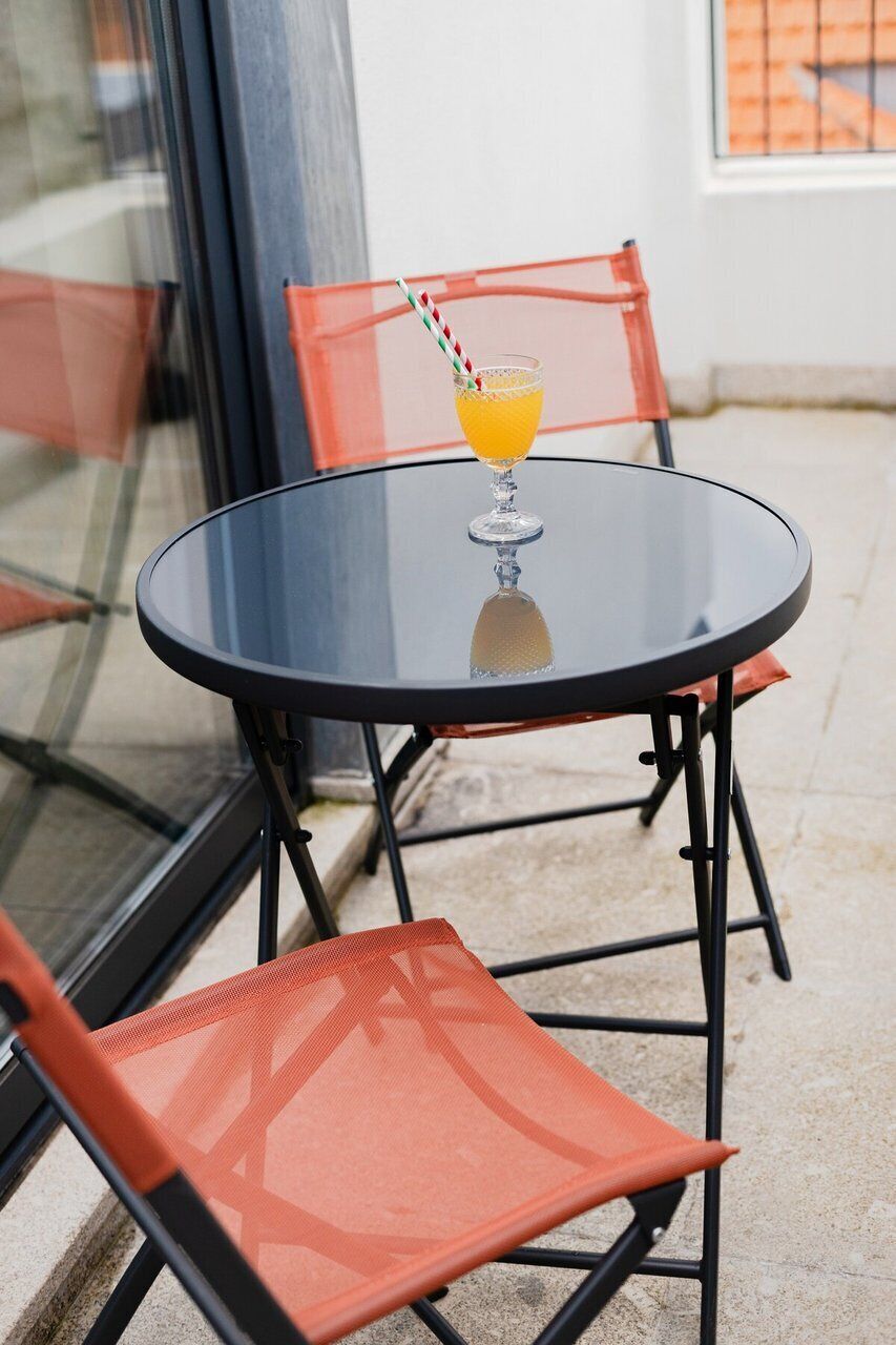 Una mesa de vidrio con un vaso de jugo de naranja y una pajita a rayas, acompañada de dos sillas de malla naranja.