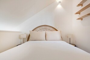 A bedroom with a bed, two bedside lamps, a woven headboard, and a slanted ceiling. - Saldanha Boho Duplex (Porto)