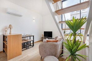 A modern living room with a sofa, a wooden side table, a TV, and a large window. - Saldanha Boho Duplex (Porto)