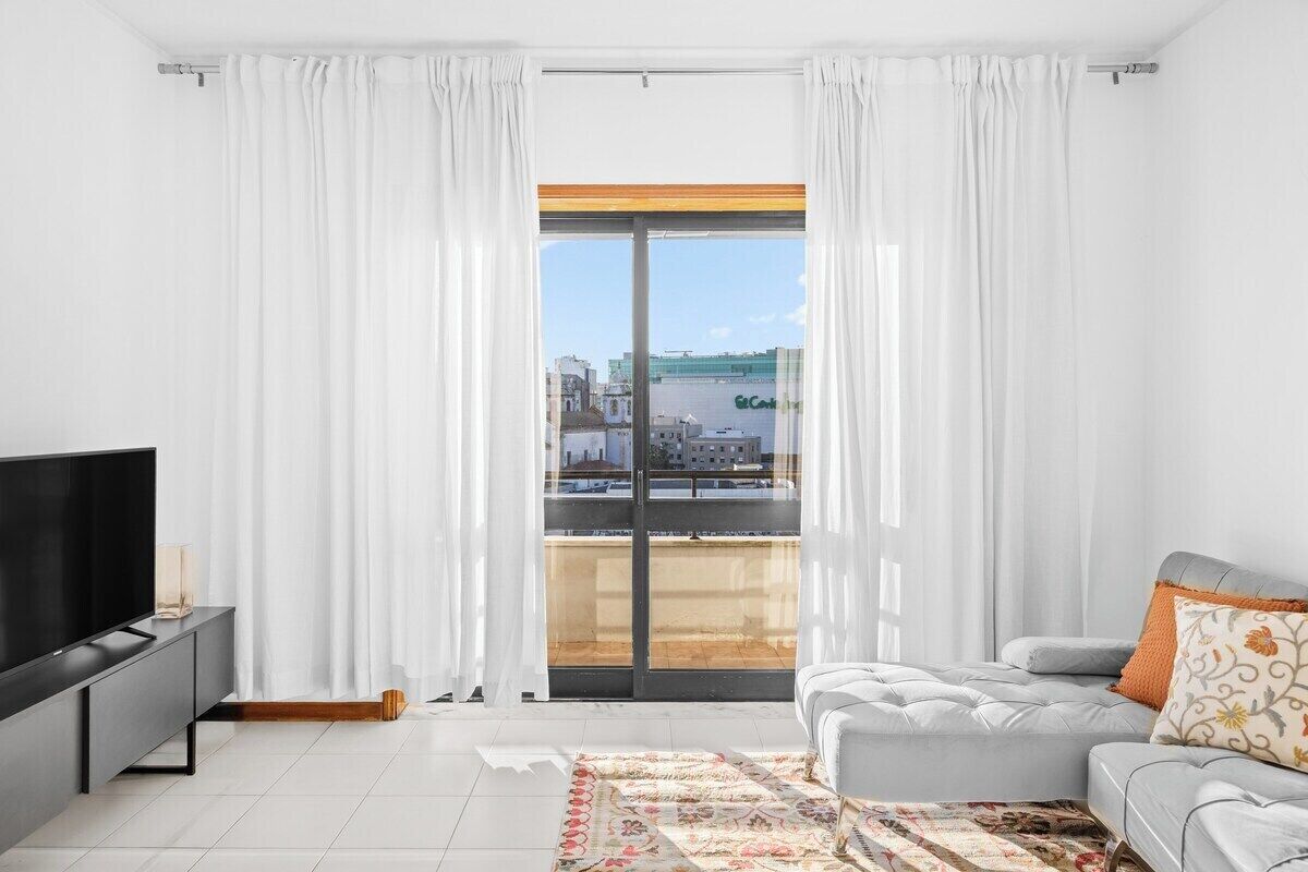 A modern living room with a large window, a flat-screen TV, a grey sofa, and a patterned rug.