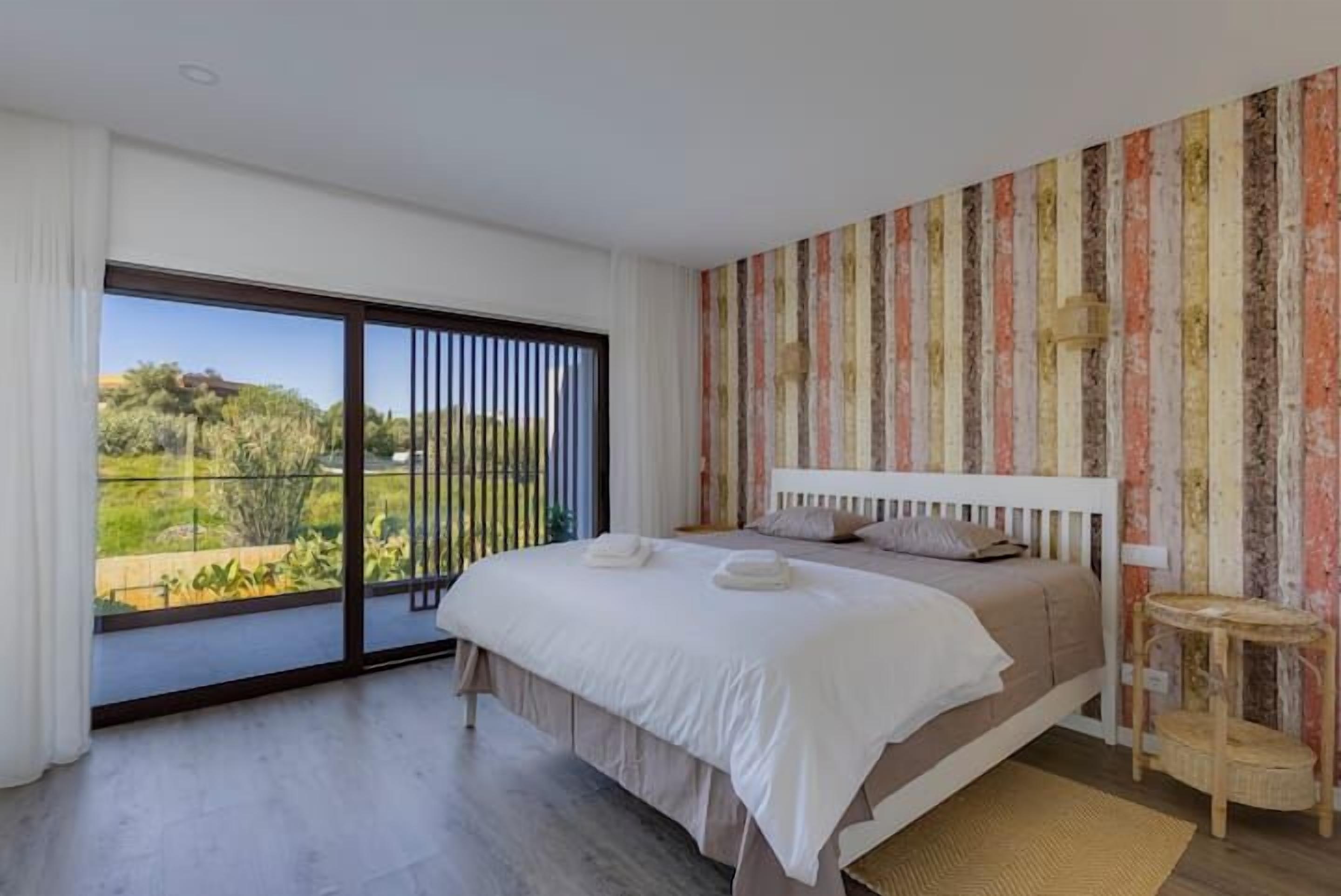 A bedroom with a large bed, a view through a sliding door, and a striped wall.