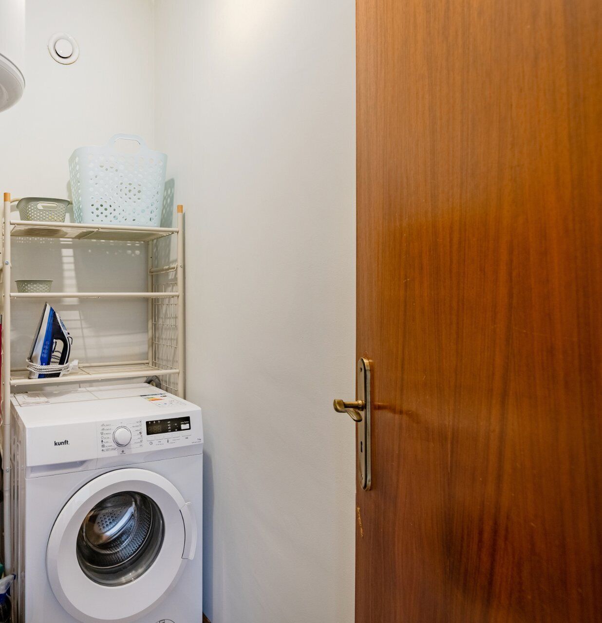 A compact laundry room with a front-loading washing machine, shelves with baskets, and a wooden door.