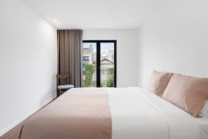 Ein modernes Hotelzimmer mit einem großen Bett, einem kleinen Beistelltisch, einem Sessel und Blick auf Gebäude durch eine Glastür.