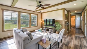 A spacious living room with a gray sofa, a wooden coffee table, and a television. There are large windows providing natural light and a view of the outdoors. - Sprawling 6-Home Yancey Ranch w/ On-Site Lake (Yancey)