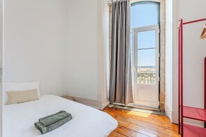 Ein Schlafzimmer mit Bett, einem Fenster mit Vorhängen, Holzboden und einem roten Metallregal.