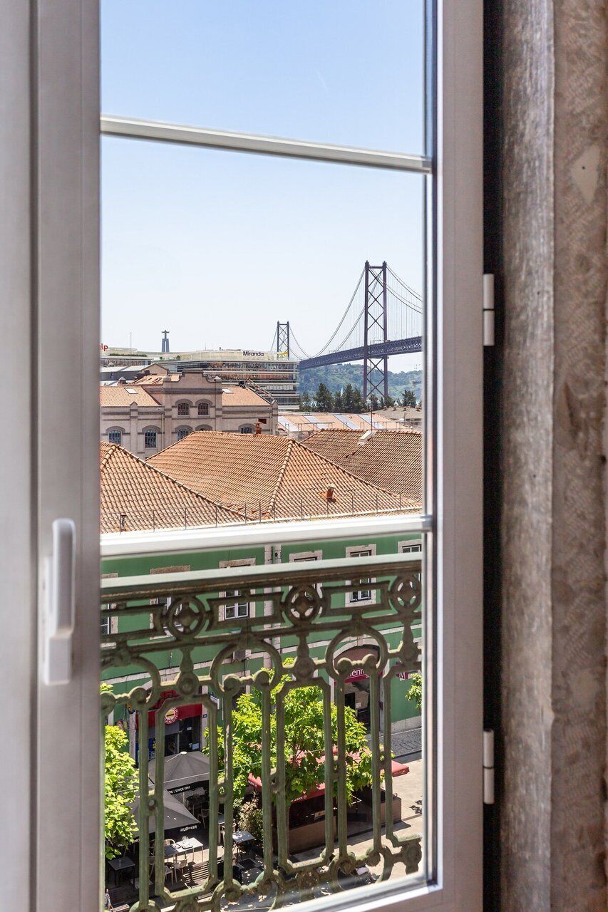 A view through a window with a metal balcony railing, a bridge, and buildings in the background.