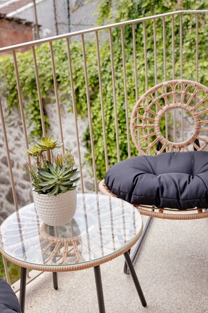 A round glass table with a woven base, a wicker chair with a cushion, and a potted plant on a balcony with a metal railing. - The Light Loft @ BoaVista Flats (Porto)