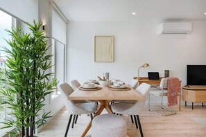 A modern dining area with a wooden table, white chairs, a potted plant, a framed picture, and a TV on a wooden stand. - The Light Loft @ BoaVista Flats (Porto)