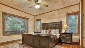 A bedroom with a wooden bed, two bedside tables, a ceiling fan, and large windows. - The Lookout | Hot Tub + Game Room + Pet Friendly (Mineral Bluff)