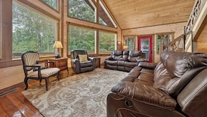 A spacious living room with large windows, a leather sofa, a chair, and a small table. - The Lookout | Hot Tub + Game Room + Pet Friendly (Mineral Bluff)