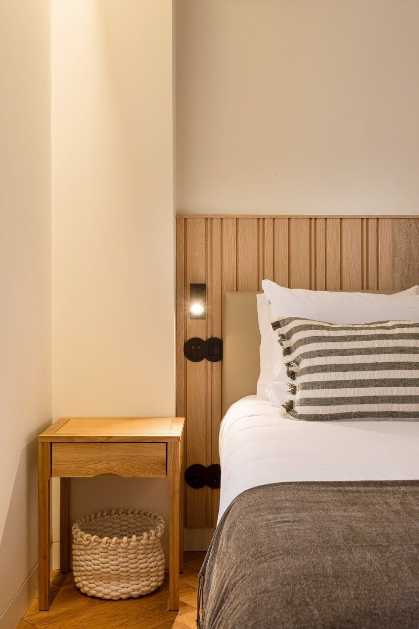 A bedroom with a wooden headboard, a bedside table with a basket, and a bed with striped pillows.